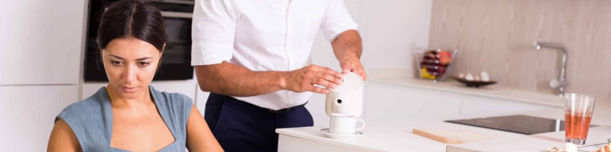 Young woman sitting with book at kitchen table while husband pouring tea