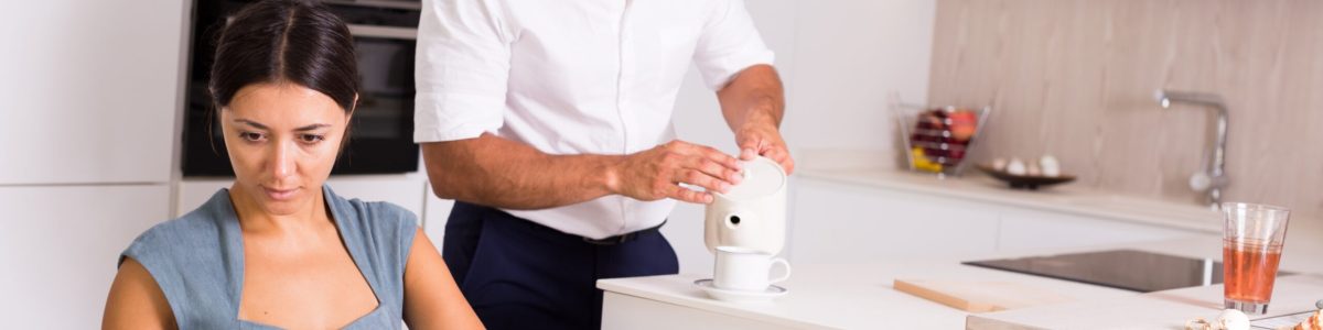 Young woman sitting with book at kitchen table while husband pouring tea
